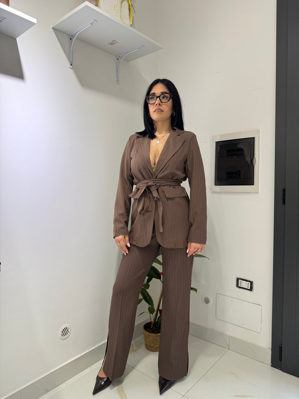 Tailleur gessato Amelie nella variante marrone cioccolato, composto da blazer con cintura annodata in vita e pantaloni a palazzo con spacco laterale sul fondo.