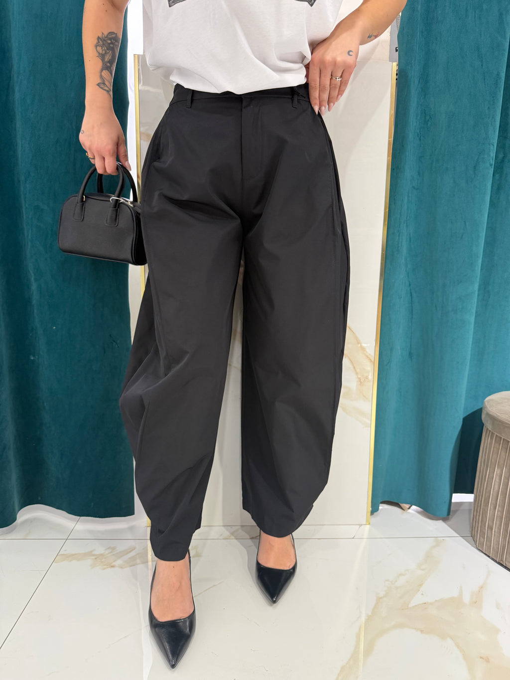 Inquadratura frontale del pantalone nero Justwest a palloncino in cotone, indossato con t-shirt bianca, décolleté a punta e borsa a mano nera.