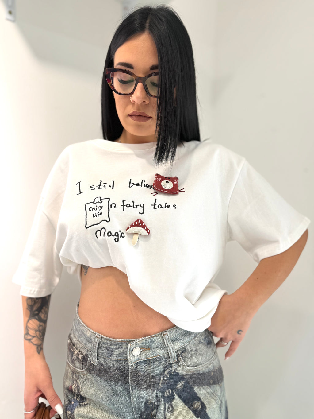 Dettaglio ravvicinato della T-shirt Over Cat bianca in cui si vedono le scritte ricamate nere e le applicazioni in tessuto imbottito in rilievo a forma di gatto e fungo.