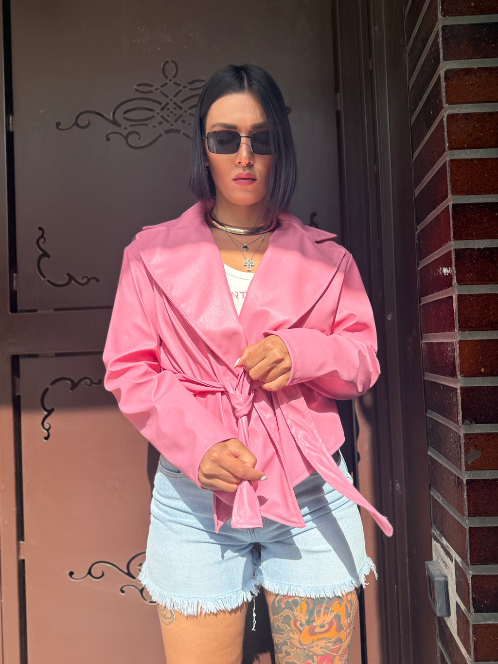 Giacca Pink corta in morbida ecopelle rosa confetto con cintura annodata in vita FAEV, indossata con top bianco e shorts in denim.