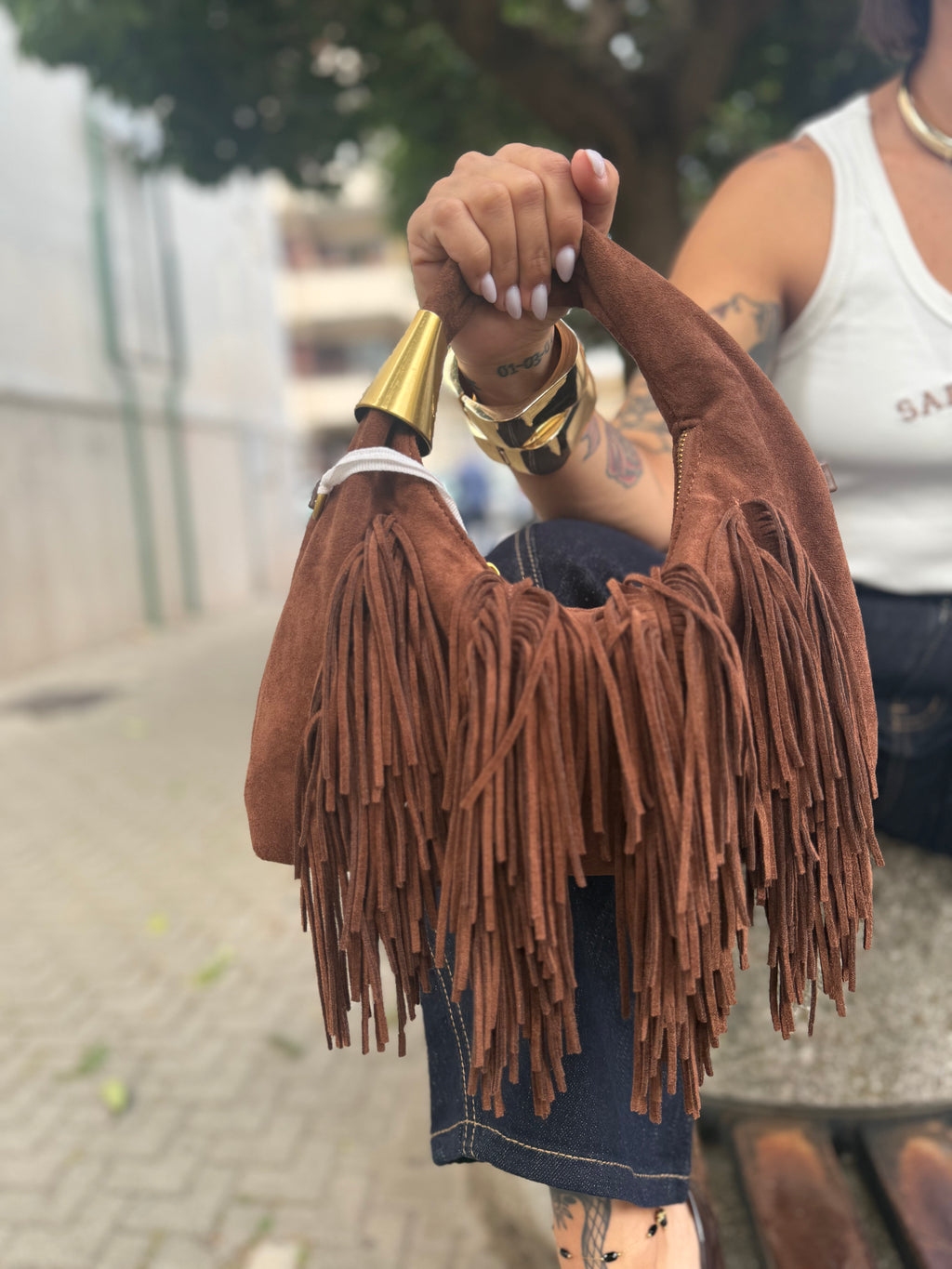 Handbag Lopez a mano in morbido scamosciato marrone castagna con lunghe frange e dettaglio metallico dorato FAEV, tenuta in mano sopra un jeans.