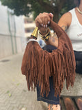 Handbag Lopez a mano in morbido scamosciato marrone castagna con lunghe frange e dettaglio metallico dorato FAEV, tenuta in mano sopra un jeans.