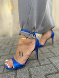Sandali Elettry in raso blu elettrico con tacco a spillo e cinturino alla caviglia FAEV, allacciati sopra un pantalone sartoriale grigio.