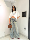 T-shirt Over Cat bianca stile oversize con scritte ricamate frontali e dettagli rialzati 3D, abbinata a jeans ampi a palazzo e scarpe con tacco bordeaux.
