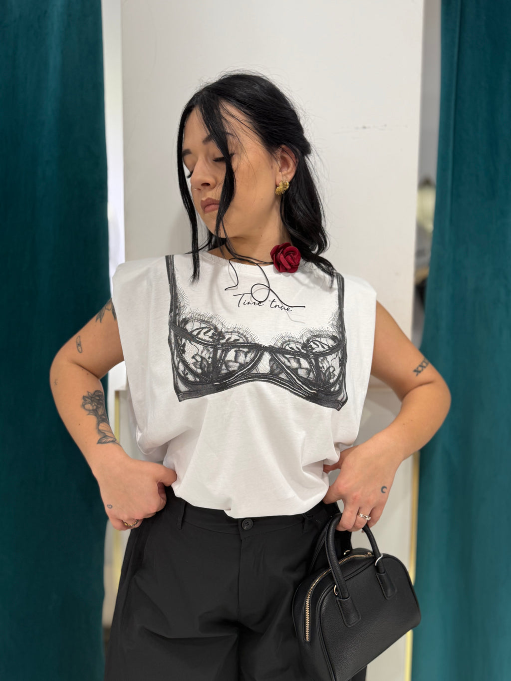 Primo piano della shirt bianca Justwest Time true in cotone con spalline imbottite e stampa frontale effetto reggiseno in pizzo nero, abbinata a pantaloni neri.