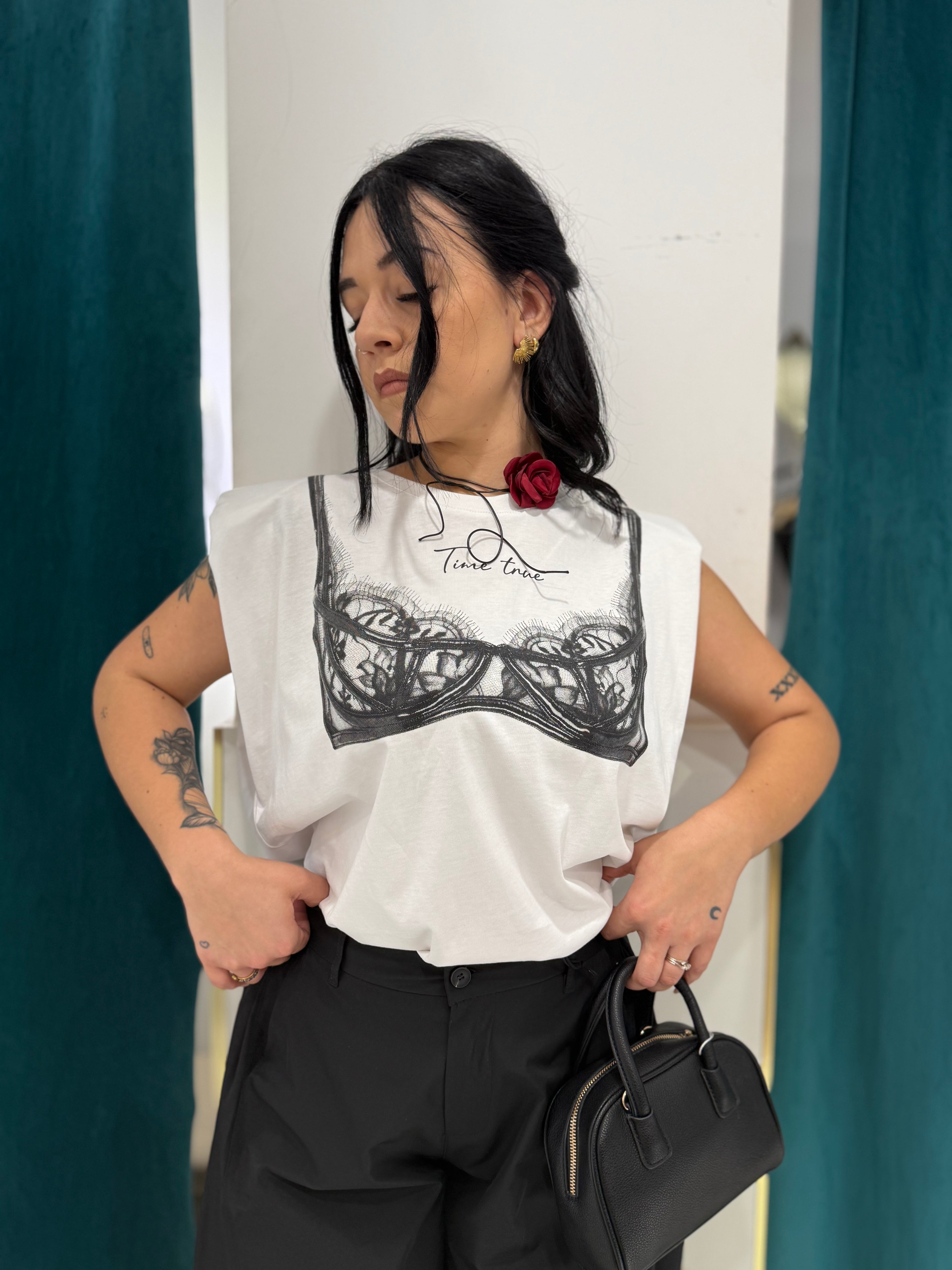 Primo piano della shirt bianca Justwest Time true in cotone con spalline imbottite e stampa frontale effetto reggiseno in pizzo nero, abbinata a pantaloni neri.