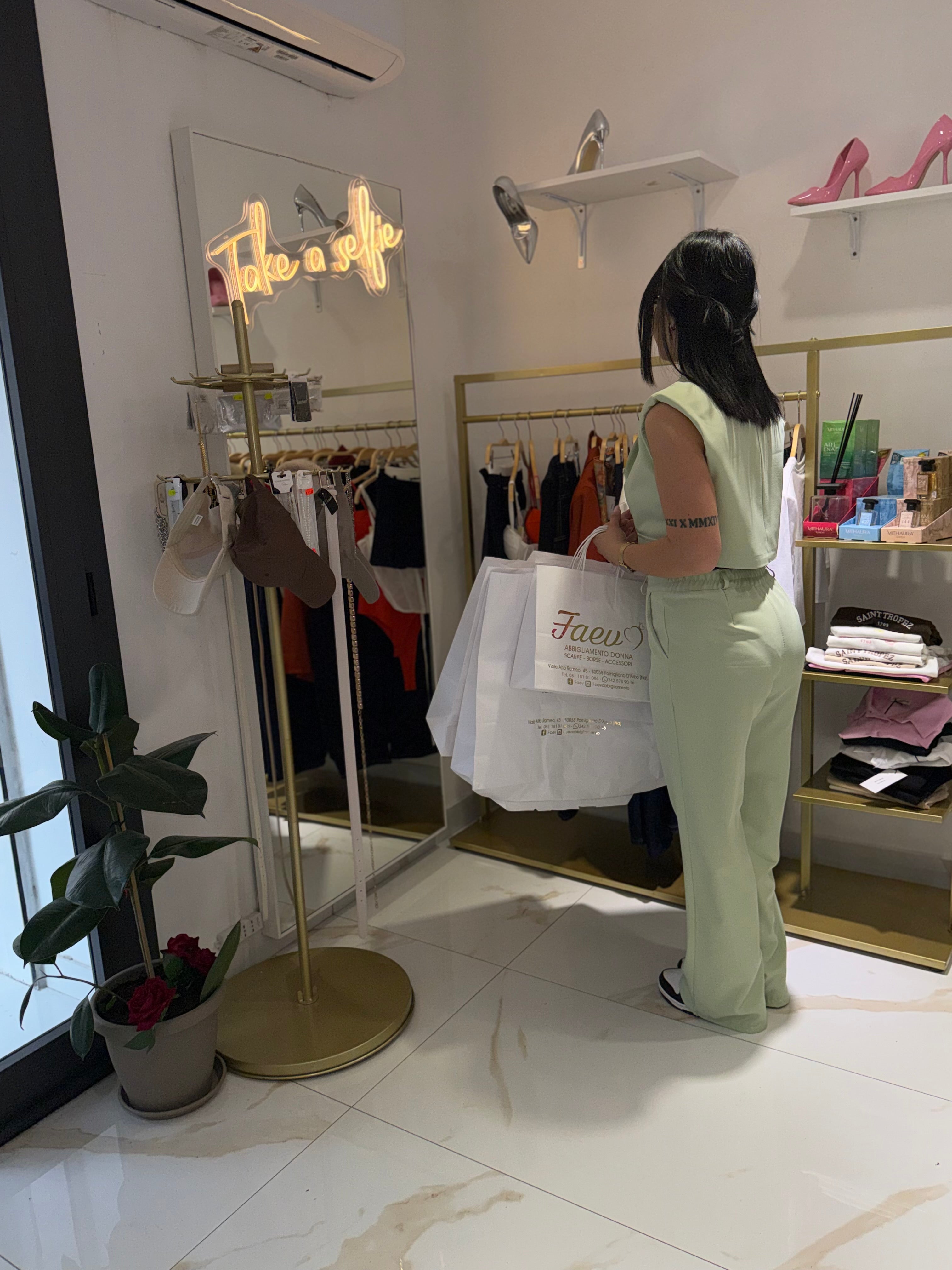 La proprietaria di FAEV Abbigliamento nella boutique a Pomigliano d'Arco con shopping bag personalizzate e didascalia al neon 'Take a selfie'.